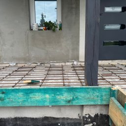 RakBud ogolno pracę remontowo budowlane. - Zbrojenie pod wylewkę na tarasie domu jednorodzinnego. Widoczne stalowe pręty, szalunek z impregnowanego drewna i fragment elewacji z oknem.