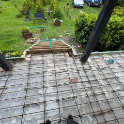 RakBud ogólne pracę remontowo budowlane. - Zbrojenie tarasu na zewnątrz z widocznymi elementami drewnianymi konstrukcji i zielonym ogrodem w tle. Widok z góry na siatkę zbrojeniową.