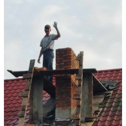 RakBud ogólne pracę remontowo budowlane. - Murarz na dachu w trakcie budowy komina z czerwonej cegły, pozdrawia. Widoczne drewniane rusztowania i fragment czerwonej dachówki. Praca na wysokości.