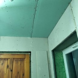 RakBud ogolno pracę remontowo budowlane. - Montaż płyt gipsowo-kartonowych na suficie i ścianach, widoczne łączenia i wkręty, surowy stan wykończenia wnętrza, drewniane drzwi i okno w tle.