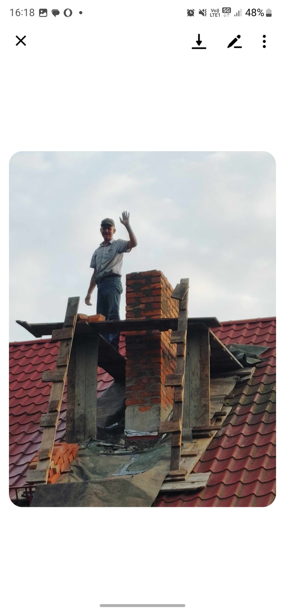 Murarz na dachu w trakcie budowy komina z czerwonej cegły, pozdrawia. Widoczne drewniane rusztowania i fragment czerwonej dachówki. Praca na wysokości.