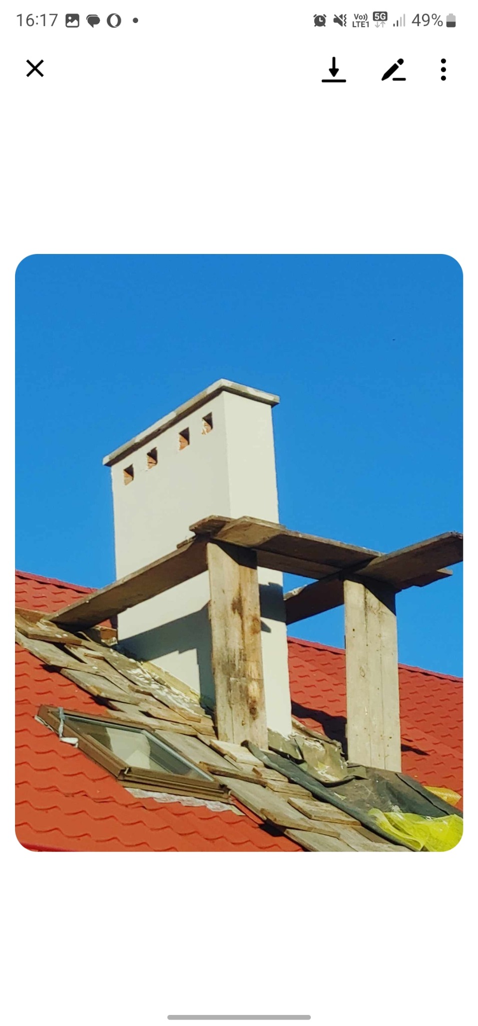 Remont dachu z kominem w Chełmie. Widoczne drewniane rusztowanie i okno dachowe. Czerwona dachówka kontrastuje z błękitnym niebem. Prace dekarskie w toku.