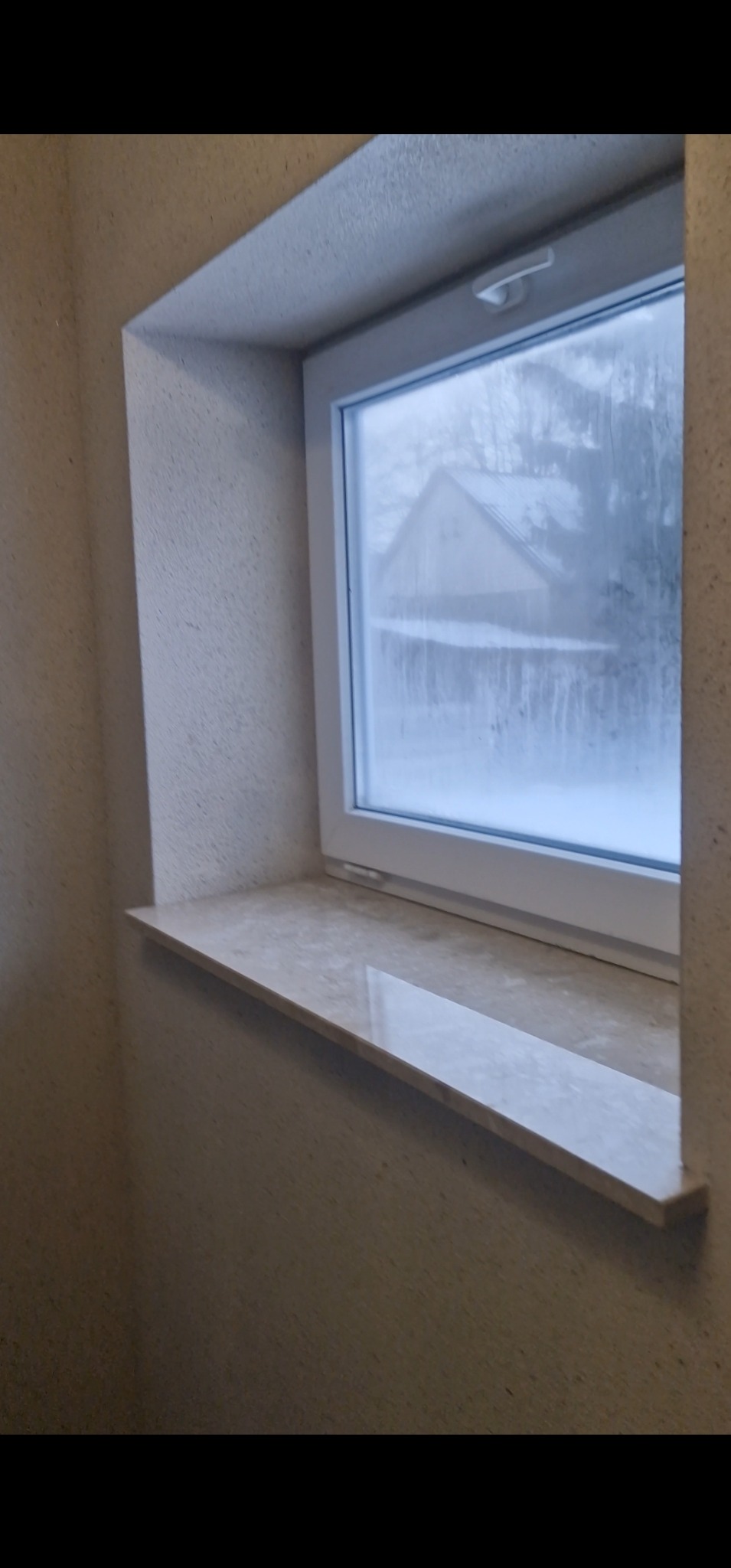 Wnętrze z oknem i marmurowym parapetem. Ściany pokryte beżowym tynkiem strukturalnym. Za oknem widok na zasypany śniegiem dom.