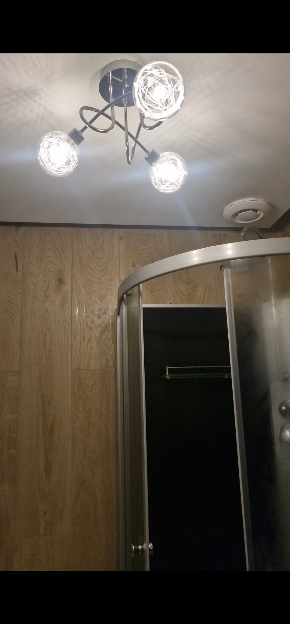 Nowoczesna lampa sufitowa z trzema żarówkami w łazience z kabiną prysznicową i panelami ściennymi imitującymi drewno. Widoczny fragment wentylacji.