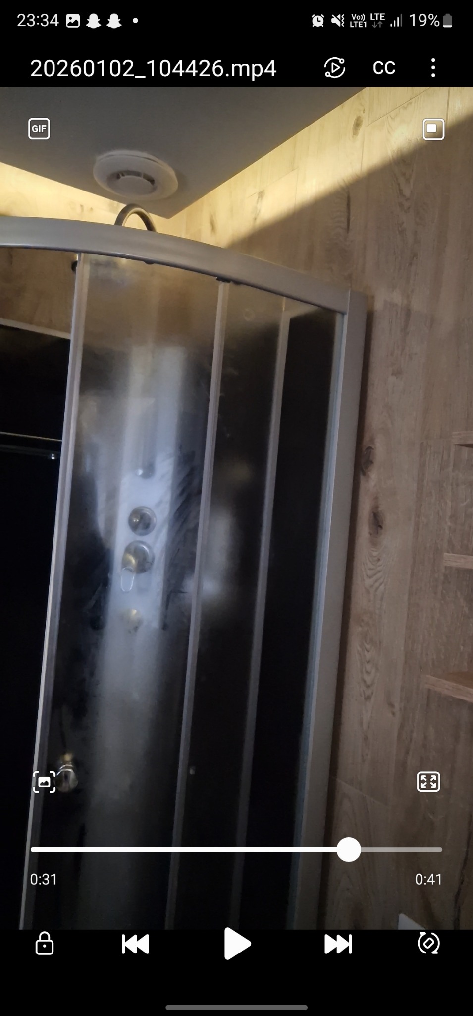 Kabina prysznicowa z przesuwnymi drzwiami, widoczna armatura i drewnopodobne panele ścienne. Ujęcie z dołu, widoczny fragment sufitu z wentylacją.