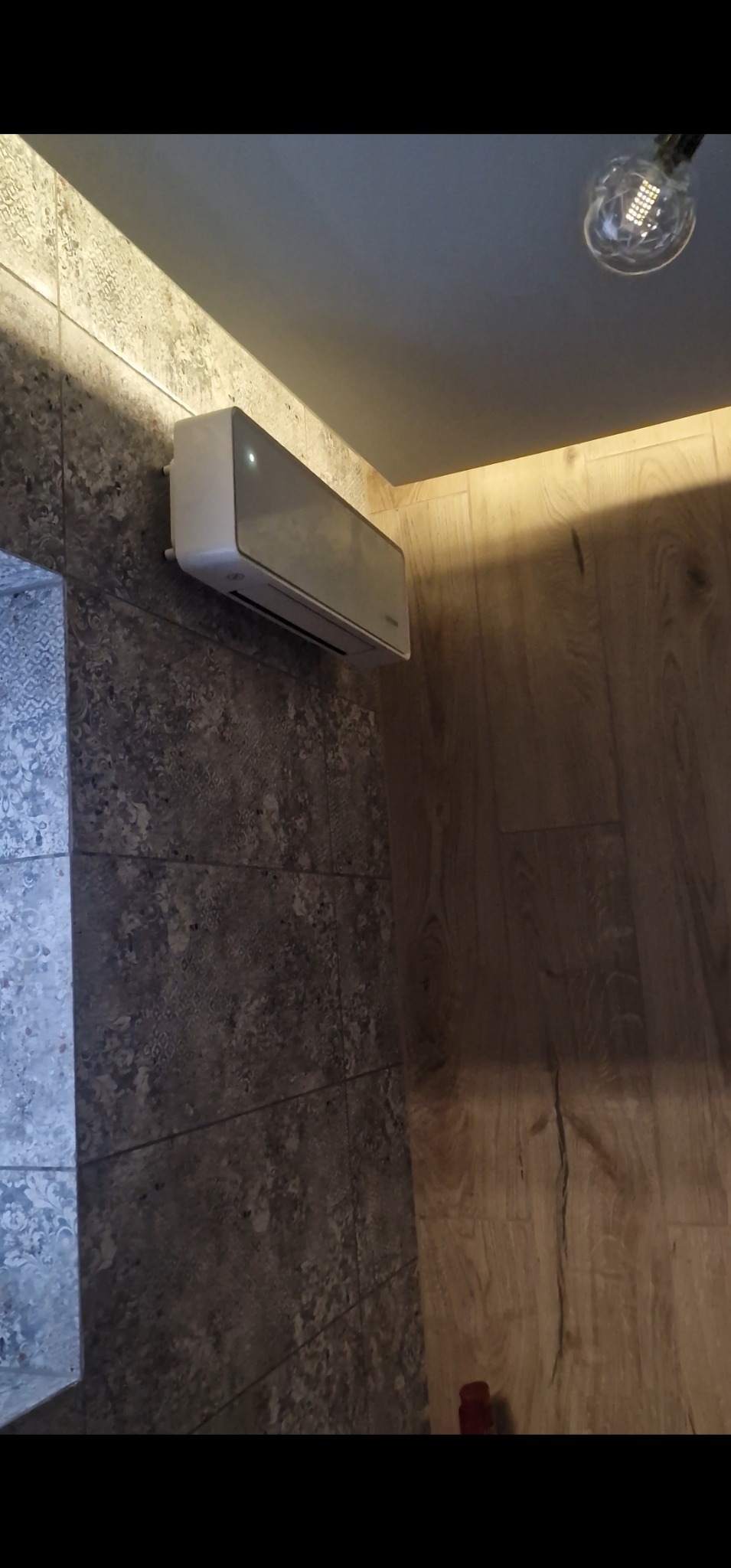 Klimatyzator na szarej, wzorzystej ścianie, obok drewniana okładzina. Widoczna lampa sufitowa. Nowoczesne wykończenie wnętrza z dbałością o detale.