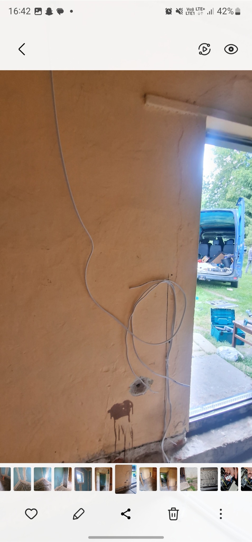 Ściana w trakcie prac elektrycznych: widoczne przewody, otwór w ścianie i ślady po instalacji. W tle częściowo widoczny samochód dostawczy z narzędziami.