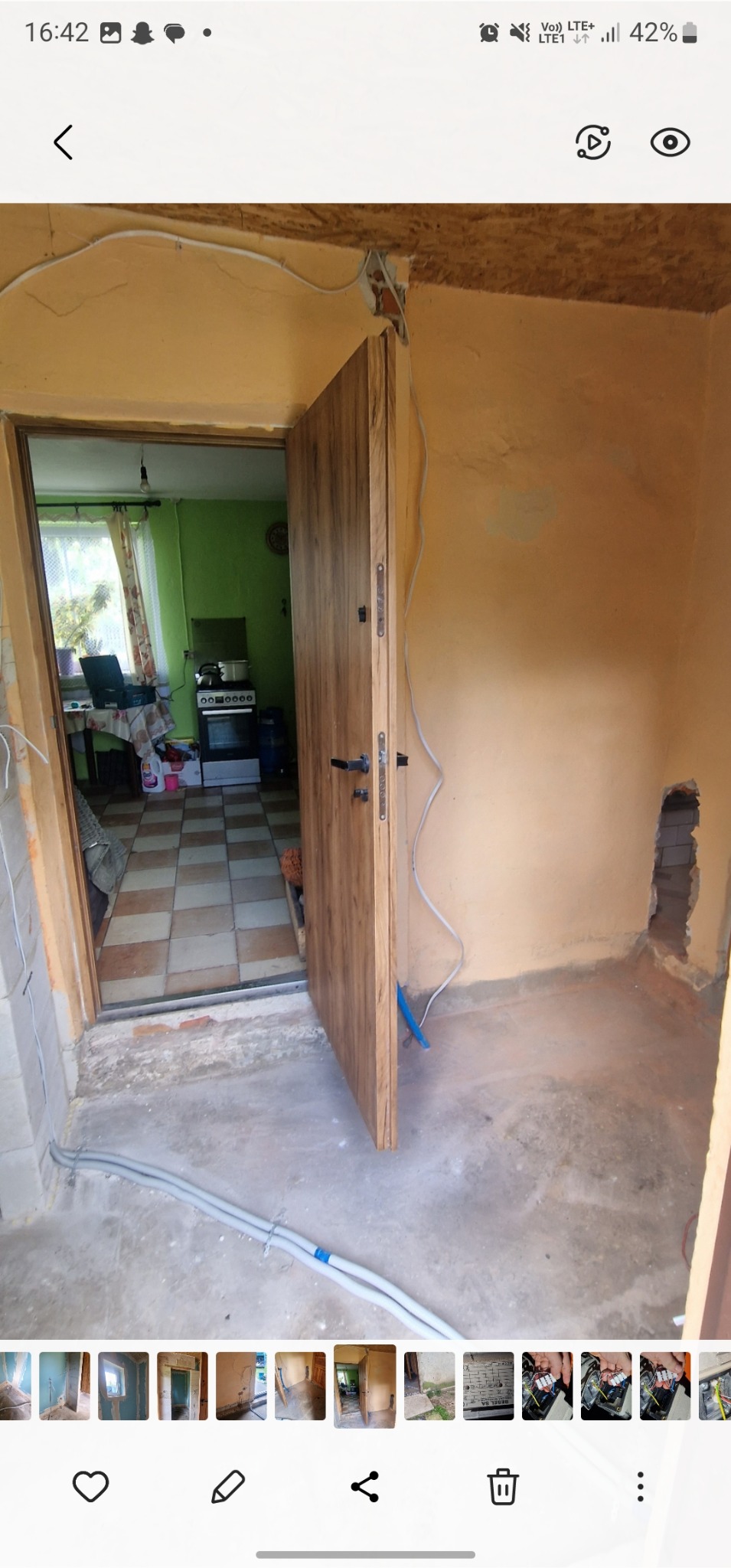 Wnętrze w trakcie remontu: widoczne kable elektryczne, surowa ściana w odcieniach beżu i otwarte drzwi z drewnianym wykończeniem prowadzące do kuchni.