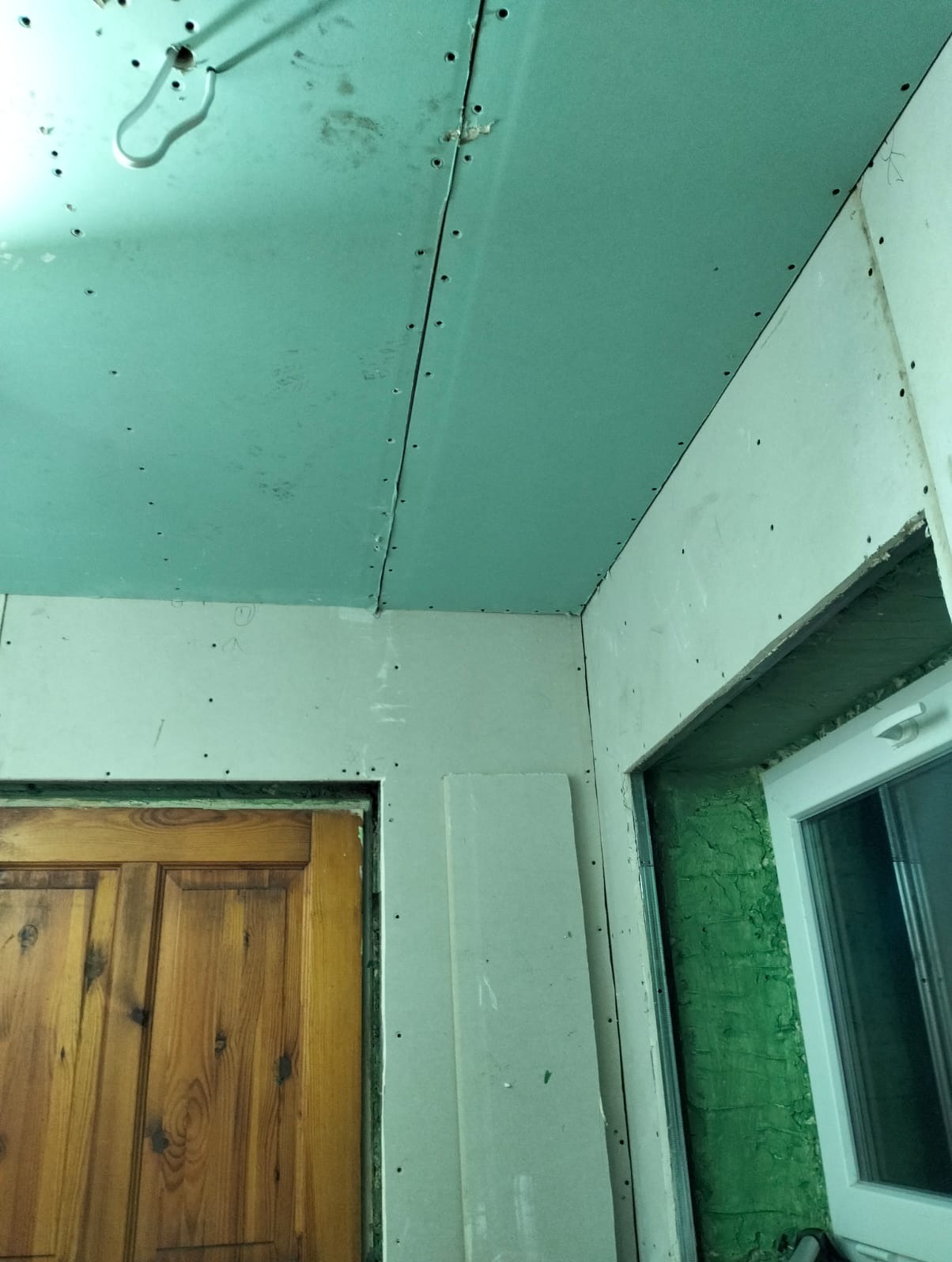 Montaż płyt gipsowo-kartonowych na suficie i ścianach, widoczne łączenia i wkręty, surowy stan wykończenia wnętrza, drewniane drzwi i okno w tle.