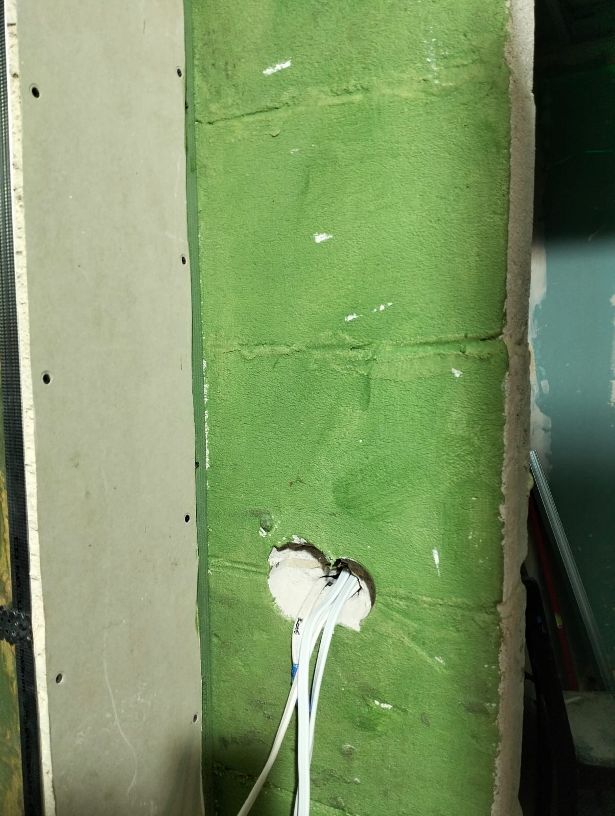 Zielona ściana z otworem na kable, obok płyta karton-gips na metalowym stelażu. Widoczne przewody elektryczne. Etap prac wykończeniowych.