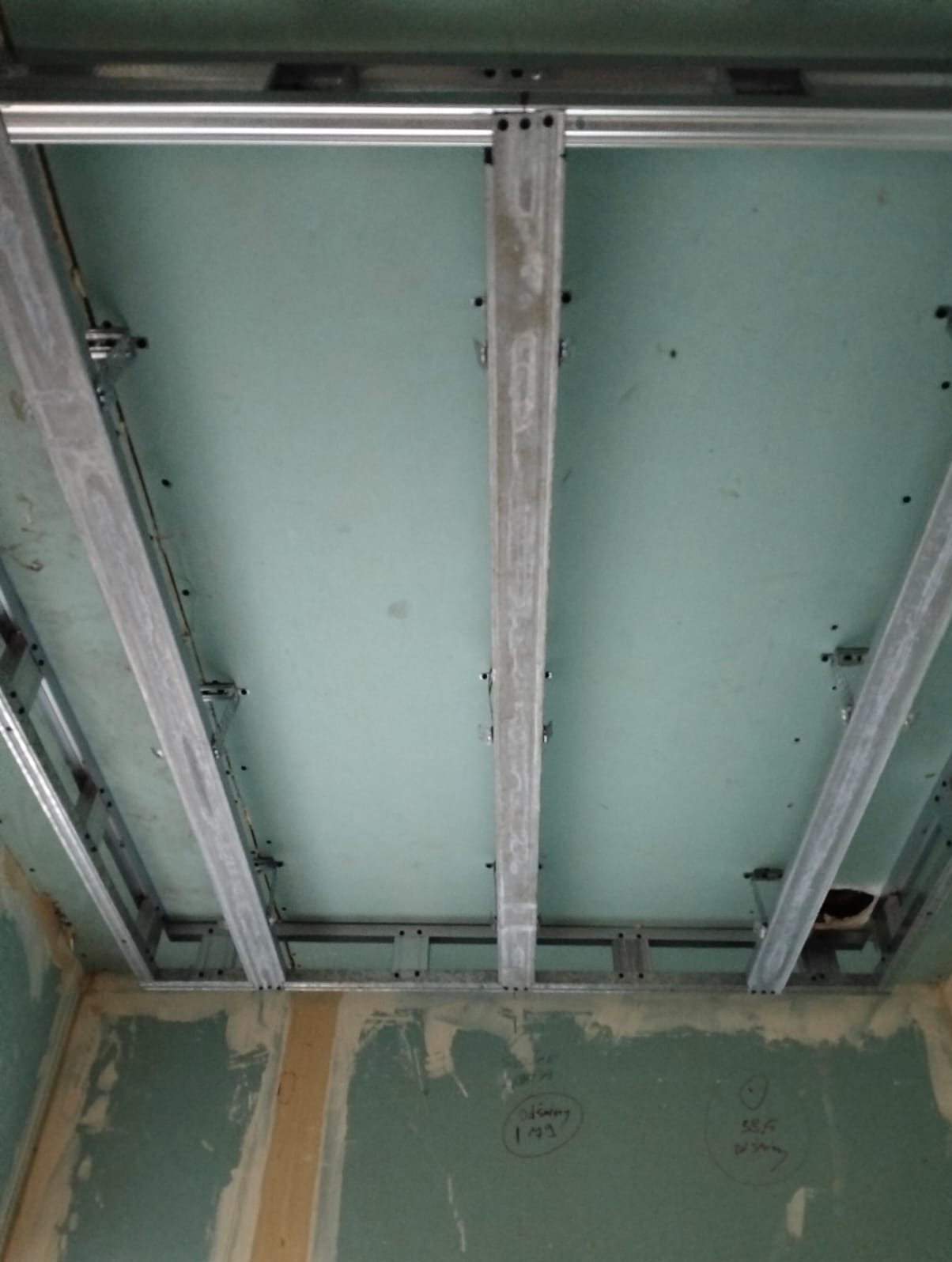 Montaż sufitu podwieszanego z płyt karton-gips: widoczne metalowe profile i płyty w kolorze zielonym, surowe ściany z widocznymi śladami prac wykończeniowych.
