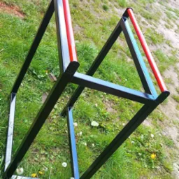 Dwie spawane, czarne, metalowe konstrukcje w kształcie trójkątów z czerwonymi poprzeczkami, leżące na trawie z widocznym samochodem dostawczym w tle.