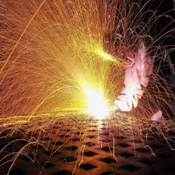 Spawalnictwo obróbka skrawaniem Michał Kuliś - Intensywne iskry spawania rozbryzgują się na ciemnym tle, oświetlając kratownicę metalową pod spodem, z widoczną dłonią spawacza w ochronnym rękawie i osłonie.