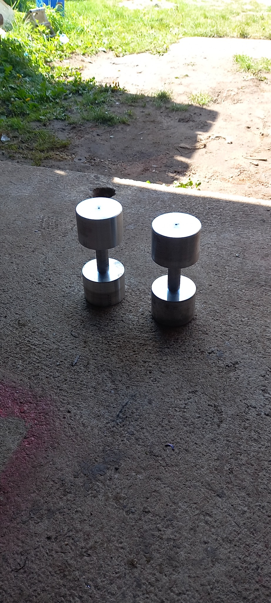 Dwie surowe, metalowe hantle o cylindrycznych obciążeniach, stojące na szorstkiej, betonowej powierzchni w półcieniu, z fragmentem trawy w tle.