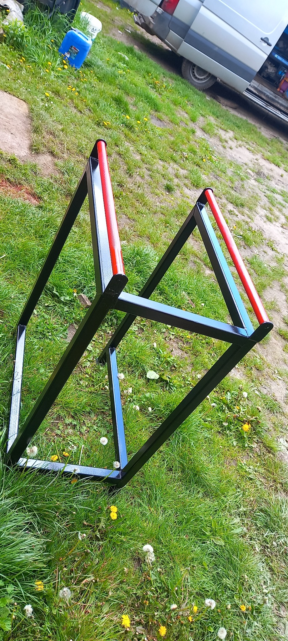 Dwie spawane, czarne, metalowe konstrukcje w kształcie trójkątów z czerwonymi poprzeczkami, leżące na trawie z widocznym samochodem dostawczym w tle.