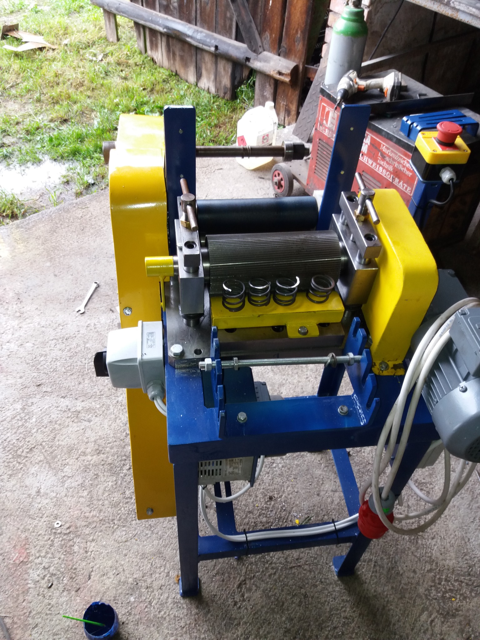 Maszyna do obróbki metali z żółto-niebieską ramą, widoczne walce i sprężyny, na podłodze leży klucz i puszka z farbą.