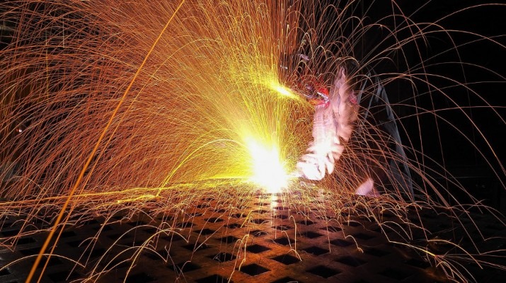 Intensywne iskry spawania rozbryzgują się na ciemnym tle, oświetlając kratownicę metalową pod spodem, z widoczną dłonią spawacza w ochronnym rękawie i osłonie.