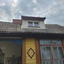 Fragment elewacji domu z oknem mansardowym, widoczne stare dachówki, drewniana weranda z wzorem rombu na żółtej desce.