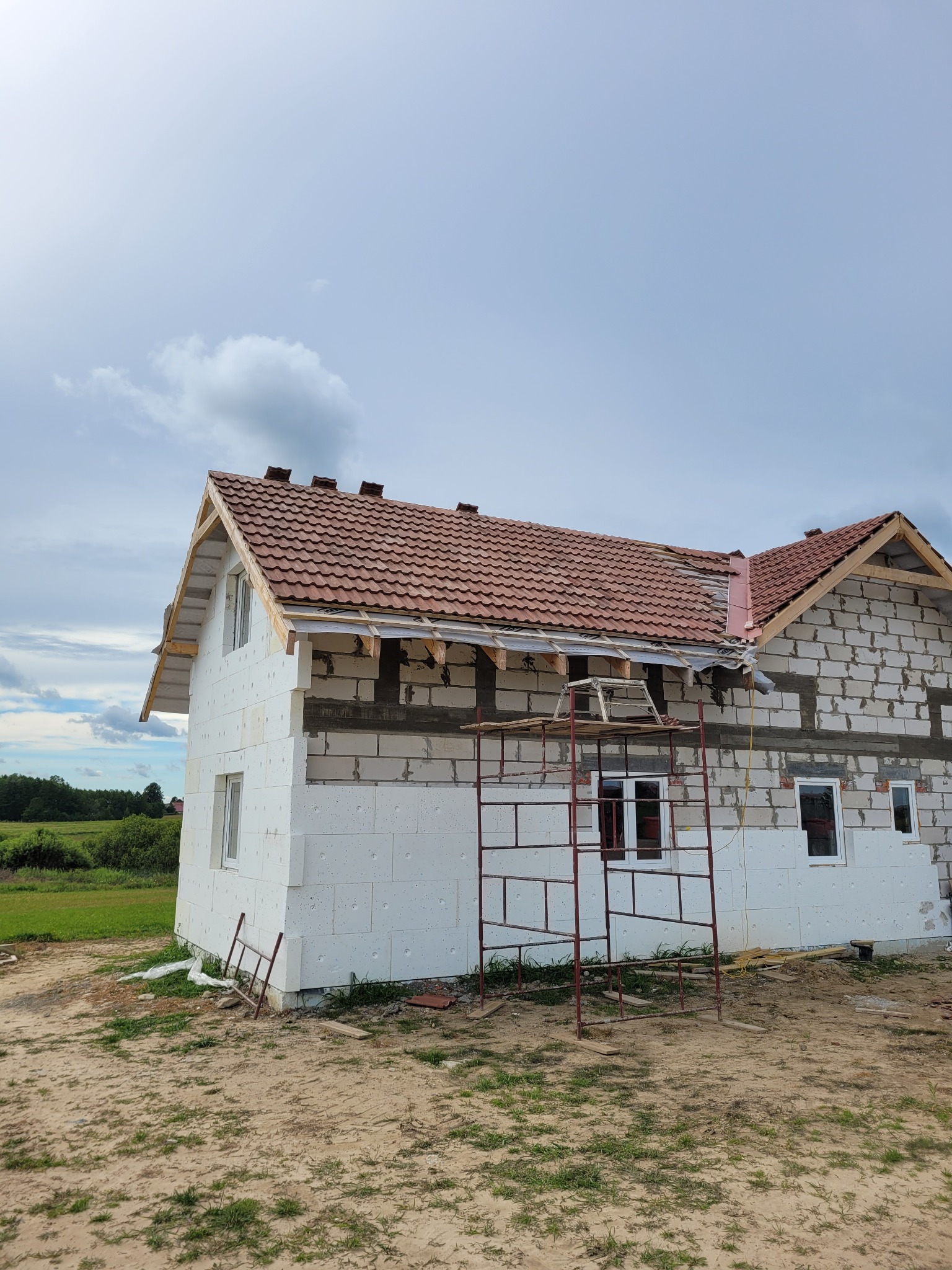Budowa domu z białych bloczków, częściowo ocieplonego styropianem, z czerwoną dachówką i rusztowaniem, widok z zewnątrz w słoneczny dzień.
