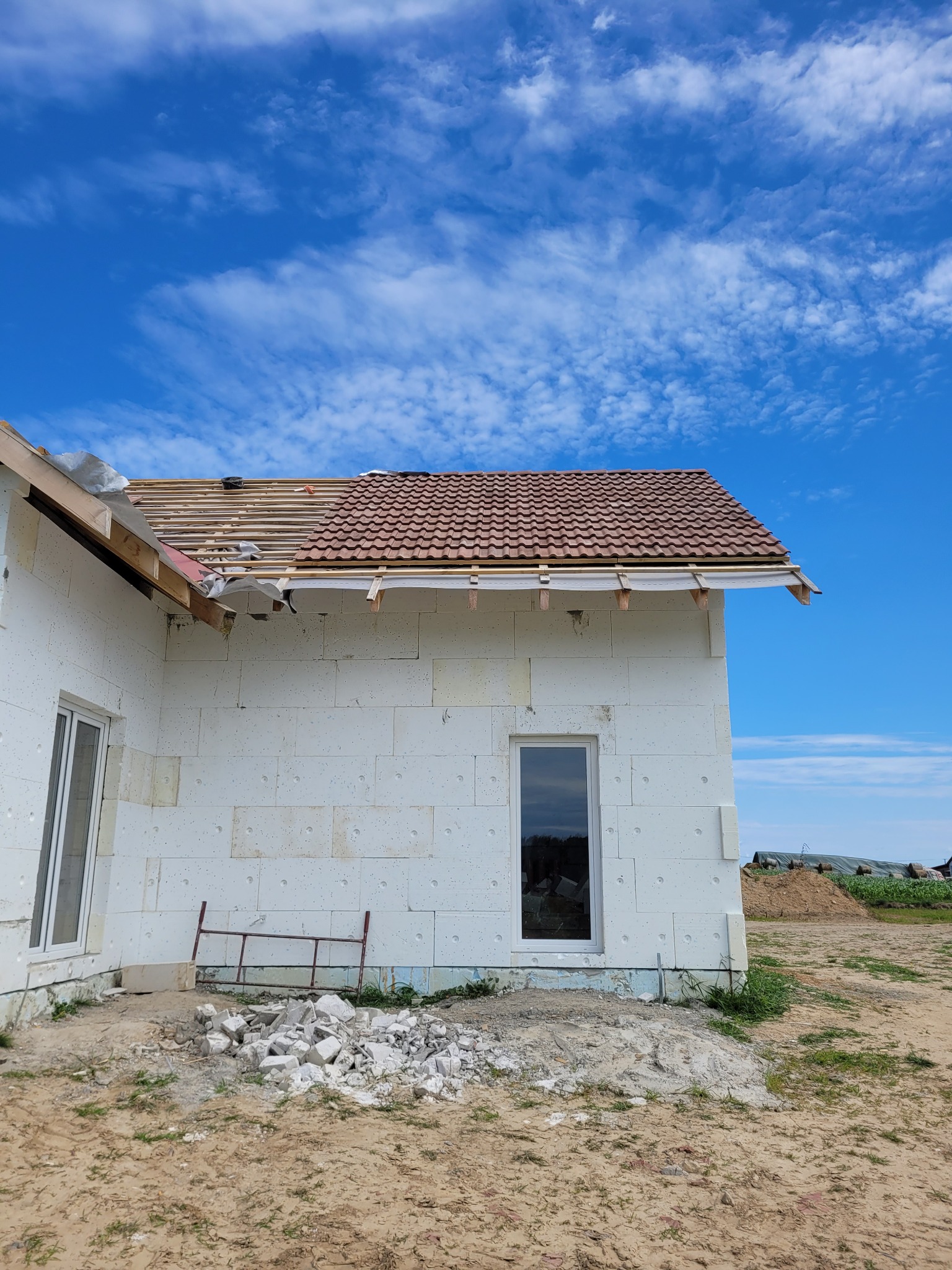 Budowa domu murowanego: ściany z białego bloczka, częściowo ułożona brązowa dachówka z widoczną konstrukcją więźby dachowej, okna, gruz i metalowa barierka na placu budowy, błękitne niebo w tle.