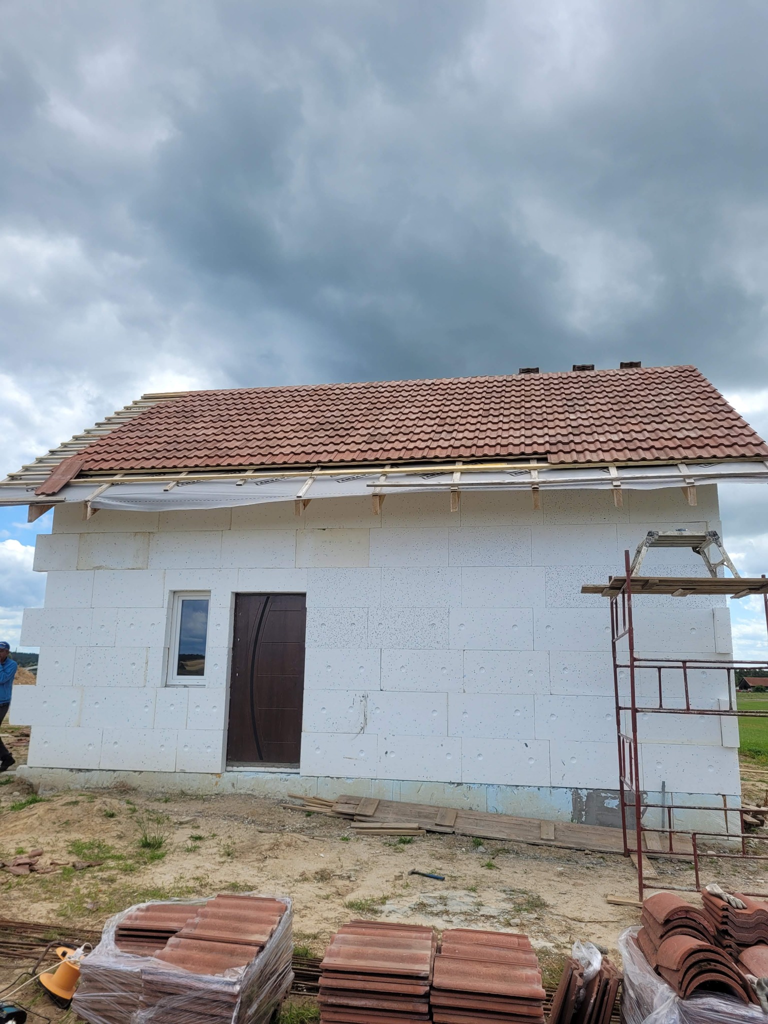 Budowa domu murowanego z białych bloczków w Pieniężnie, widoczne przygotowane dachówki i rusztowanie, częściowo ułożony dach.