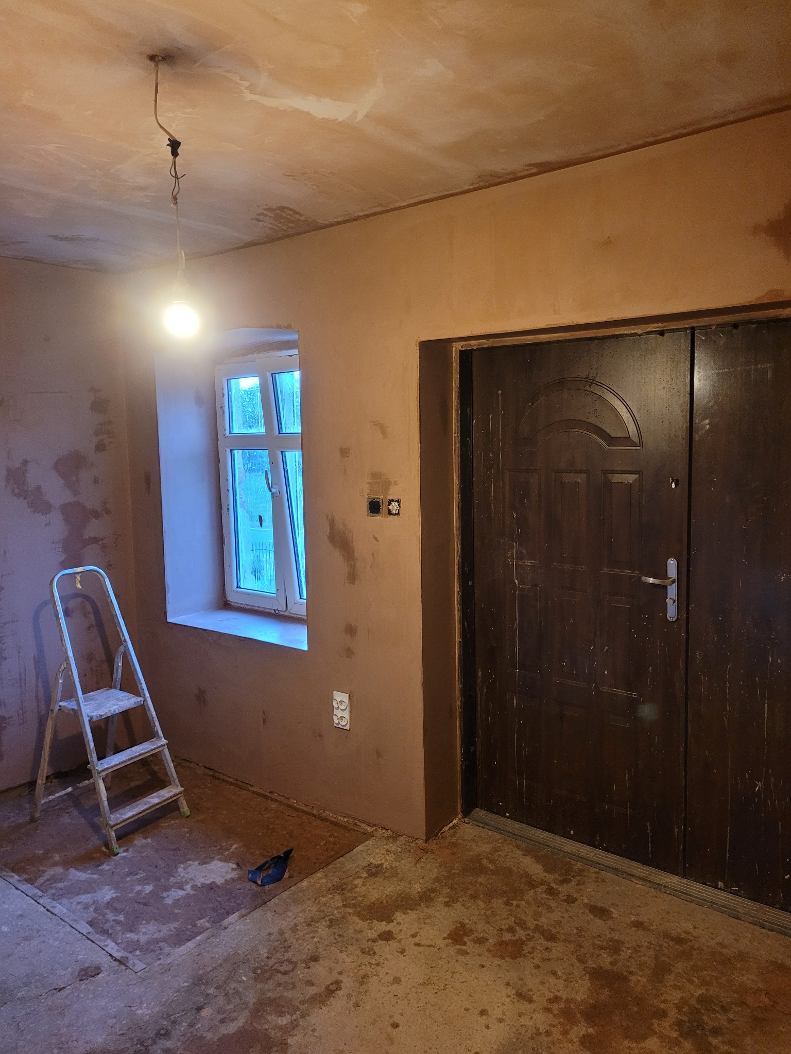 Pokój w trakcie tynkowania ścian, widoczna drabina, okno z widokiem na zewnątrz, surowy beton na podłodze i wisząca żarówka.