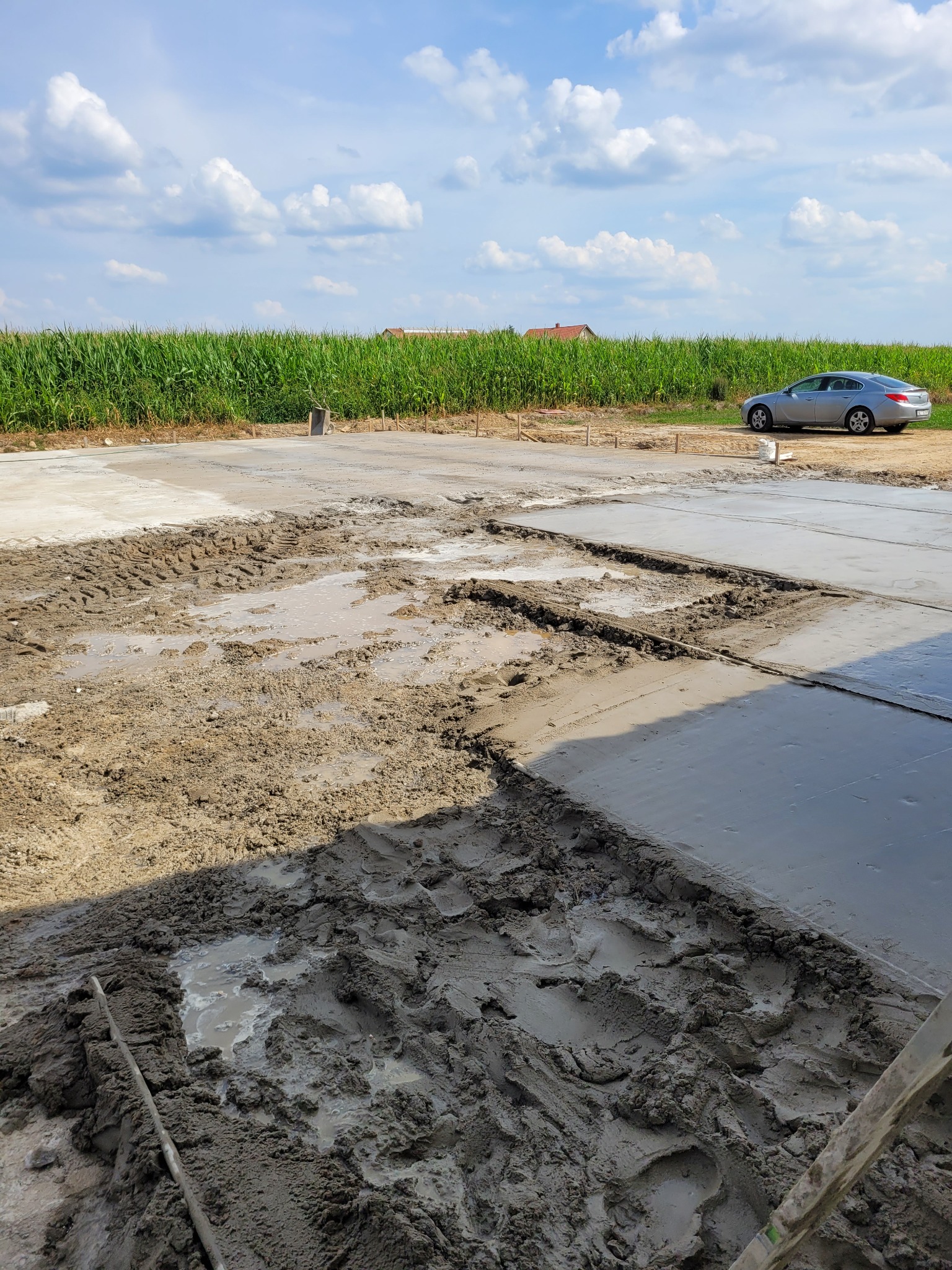 Wylany fundament domu w trakcie prac, widoczne ślady opon i błoto, w tle pole kukurydzy i samochód osobowy.