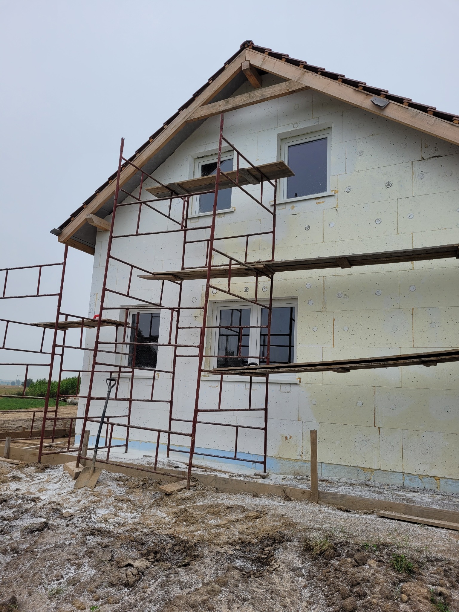 Budowa domu murowanego: ściany z białego styropianu, rusztowanie metalowe, widoczne okna i elementy konstrukcji dachu, szpadel oparty o rusztowanie, teren wokół domu w trakcie prac ziemnych.