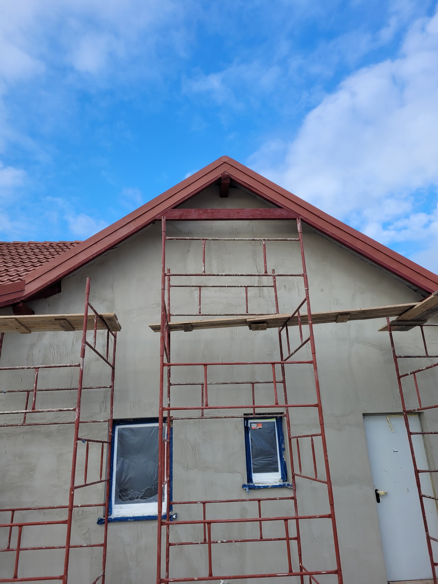 Dom w trakcie tynkowania, widoczne rusztowania, okna zabezpieczone folią, czerwona dachówka i fragment błękitnego nieba.