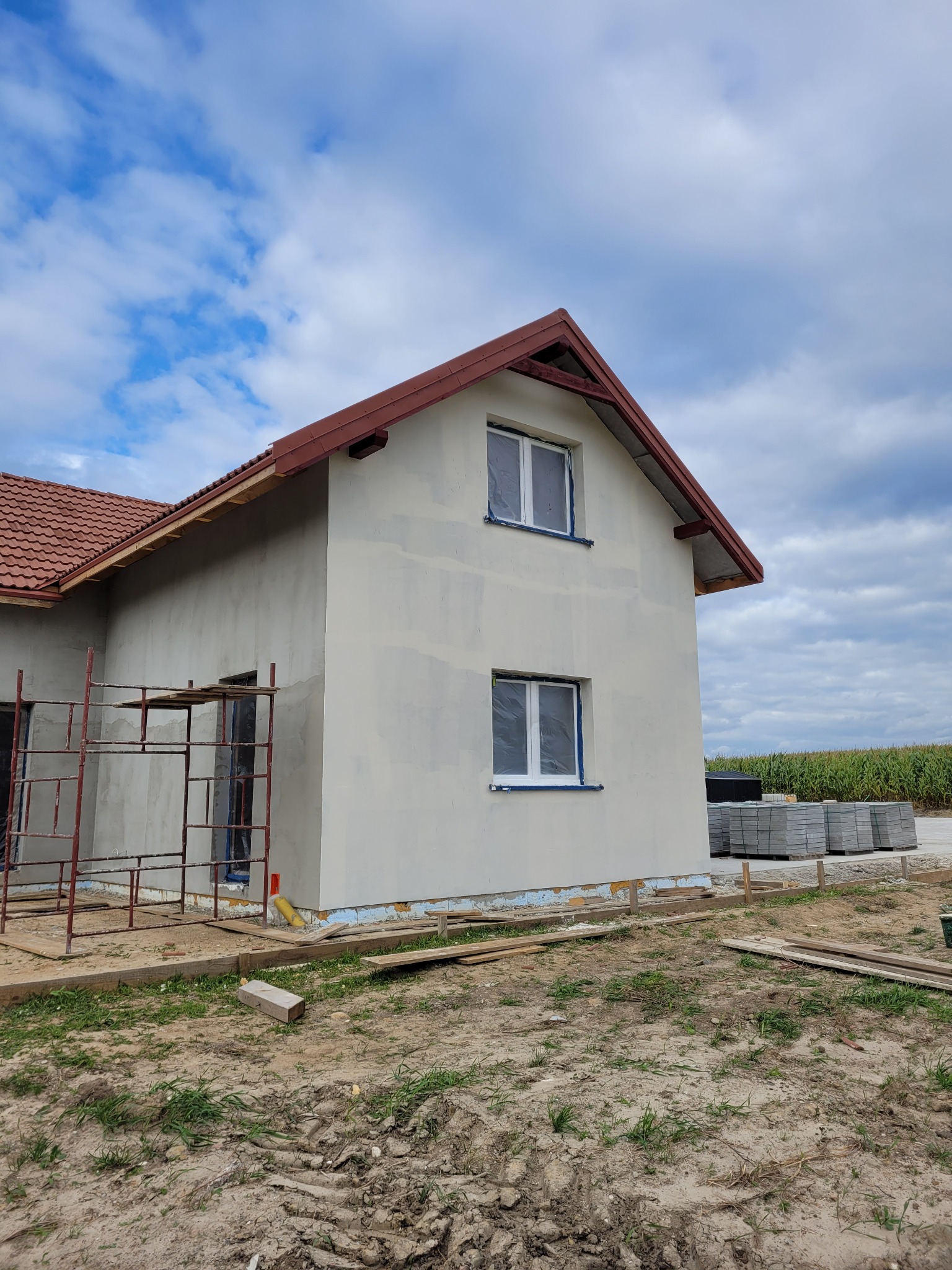 Surowy, dwukondygnacyjny dom z czerwoną dachówką w trakcie budowy, z częściowo otynkowaną elewacją i rusztowaniem z przodu, okna zabezpieczone folią, z widocznymi materiałami budowlanymi obok.