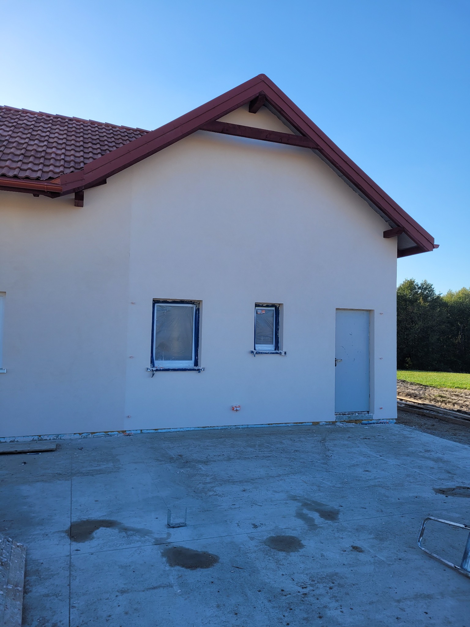 Niedokończony dom murowany z czerwoną dachówką, okna zabezpieczone folią, surowy betonowy podkład i widoczne ślady po budowie.