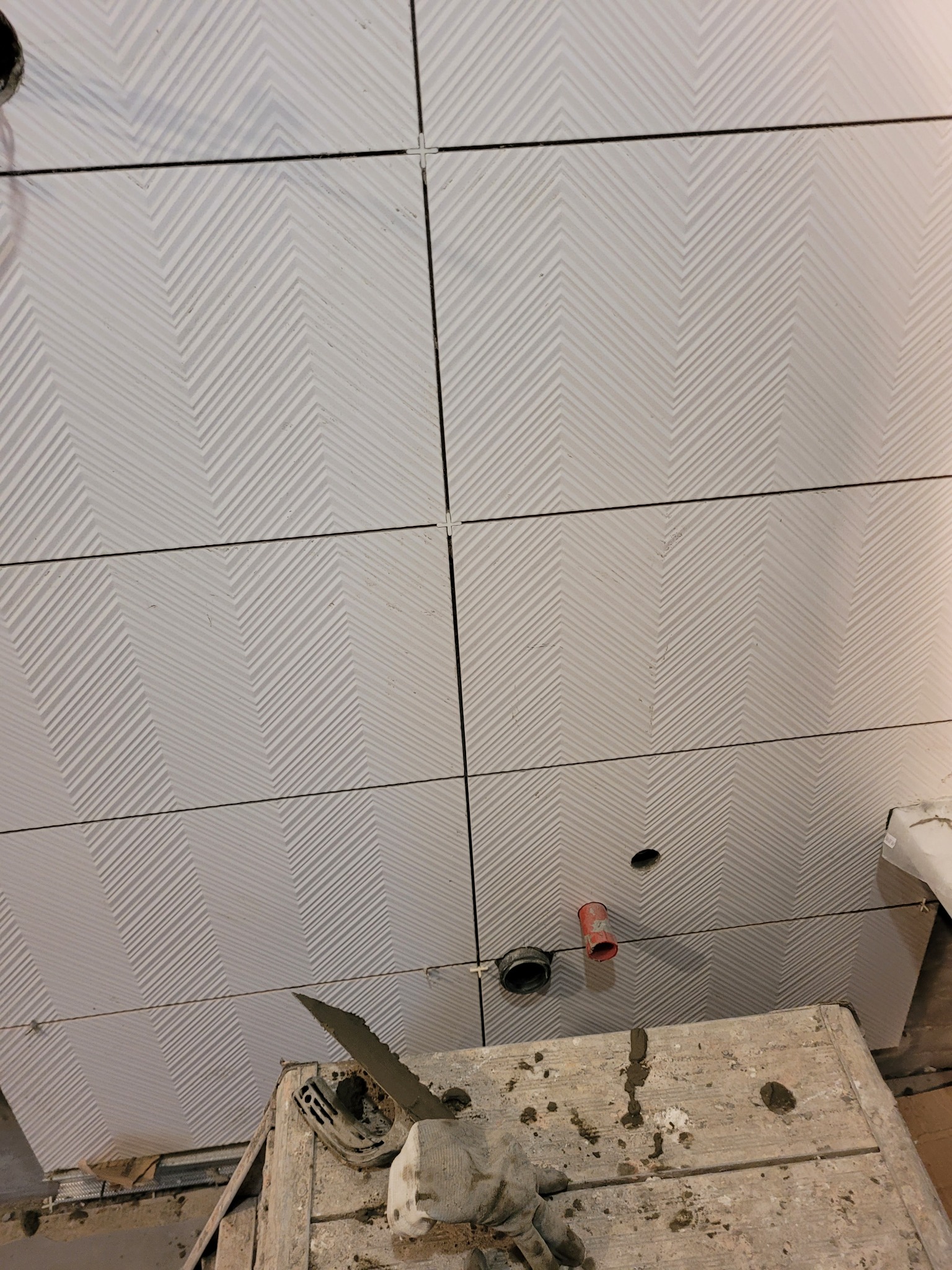 Montaż płytek ściennych z wzorem w jodełkę, widoczna drabina murarska z narzędziami i zaprawą, detale instalacji elektrycznej.