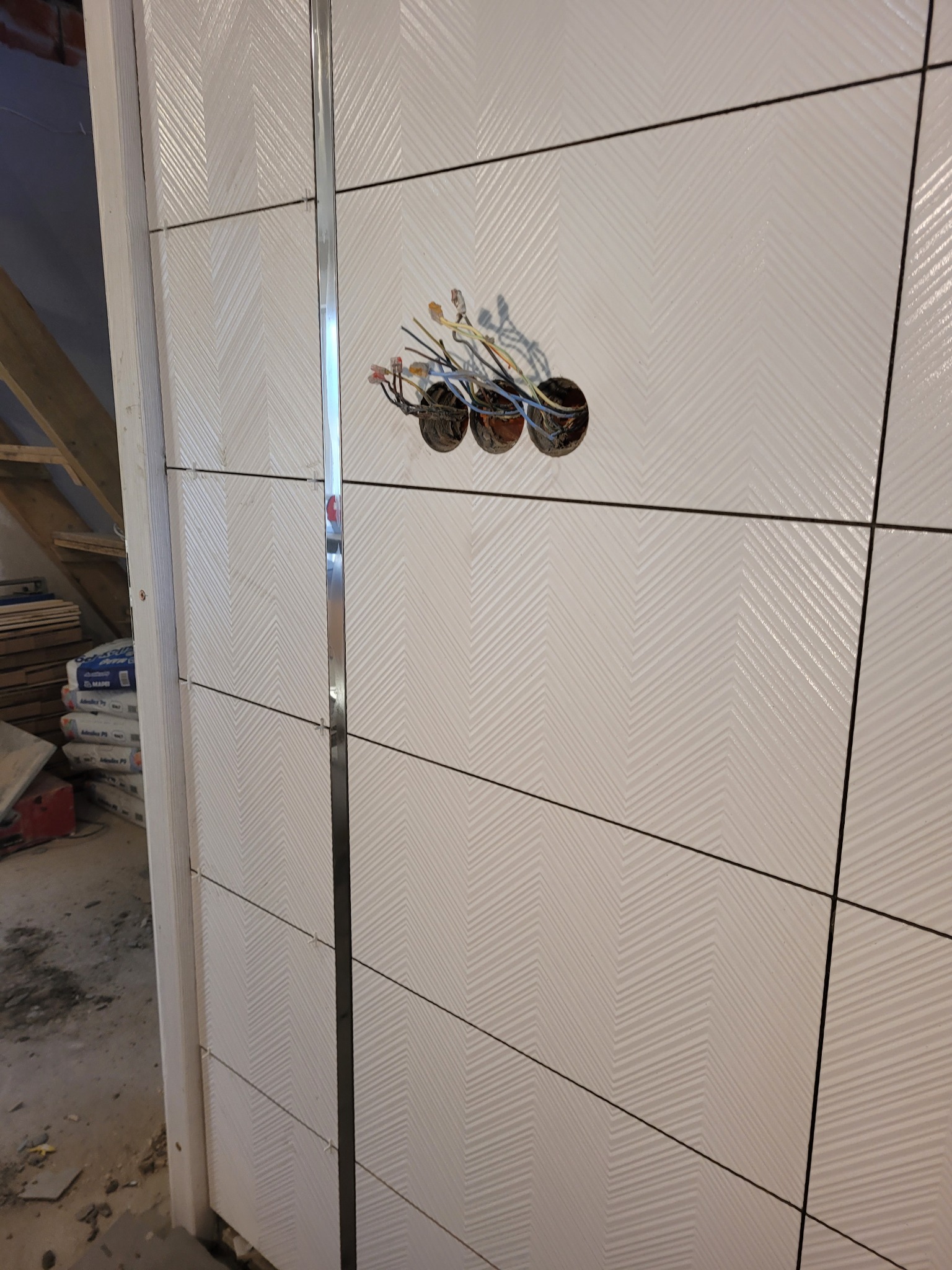 Ściana wyłożona białymi płytkami z wzorem w jodełkę, z widocznymi trzema otworami na puszki elektryczne i wystającymi przewodami, oddzielona metalową listwą.