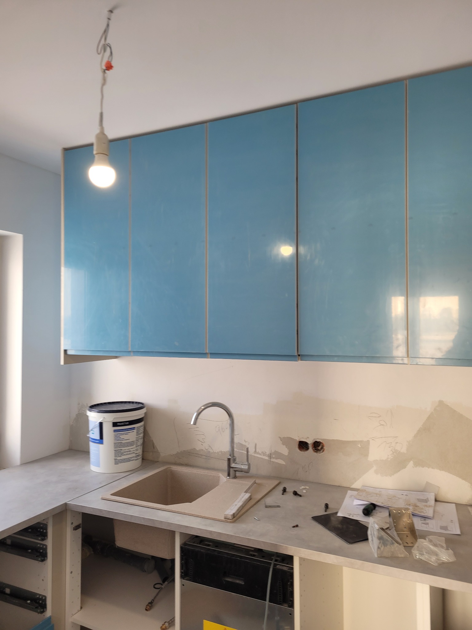 Niedokończona kuchnia z jasnoniebieskimi szafkami wiszącymi, beżowym zlewem, szpachlowaną ścianą z widocznymi gniazdkami i wiszącą żarówką