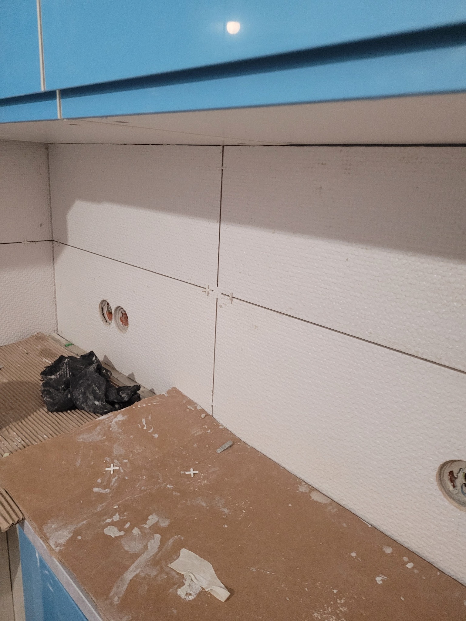 Fragment ściany w trakcie układania białych, teksturowanych płytek, widoczne krzyżyki dystansowe, otwory pod gniazdka elektryczne i fragment niebieskich szafek kuchennych.