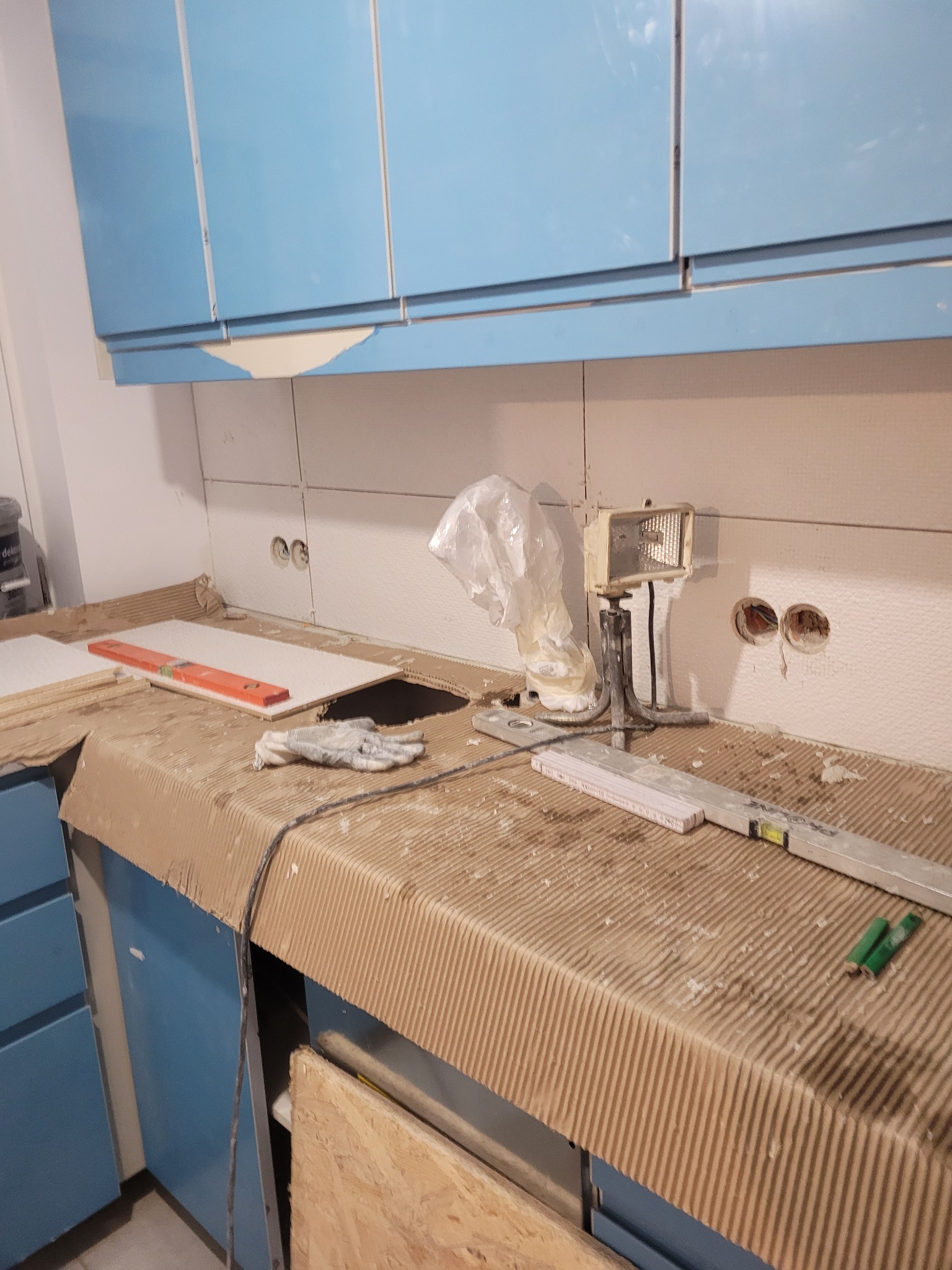 Niedokończona kuchnia: białe płytki ścienne z widocznymi puszkami elektrycznymi, blat zabezpieczony kartonem, poziomica, rękawice robocze i lampa budowlana.