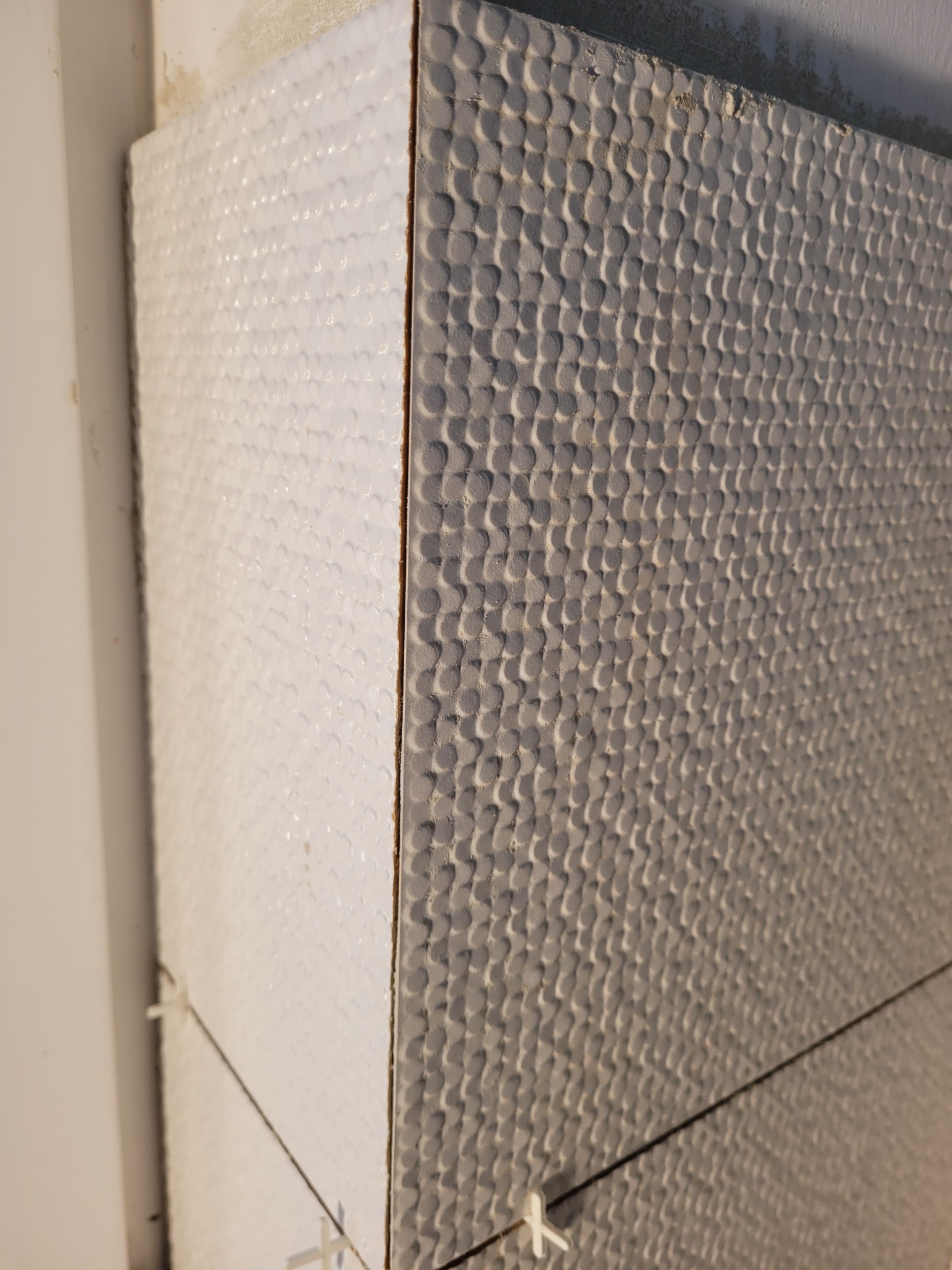 Ułożone płytki ścienne z wyraźną fakturą, krzyżyki dystansowe widoczne w spoinach, surowy stan wykończenia ściany w tle.