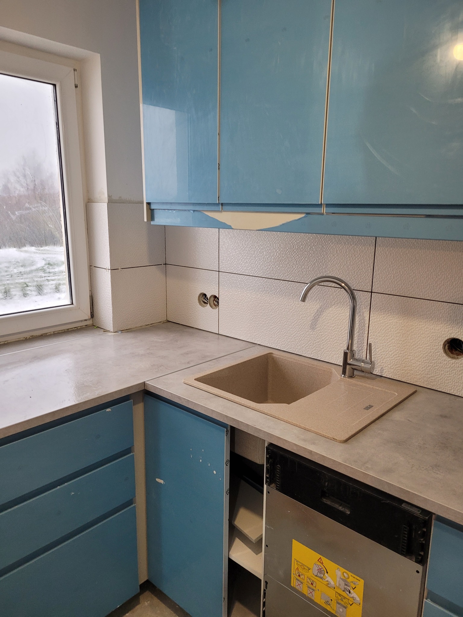 Fragment kuchni w trakcie remontu: niebieskie szafki z uszkodzonym lakierem, beżowy zlew i bateria, białe płytki z fakturą, widoczne gniazdka i niedokończona zabudowa zmywarki.