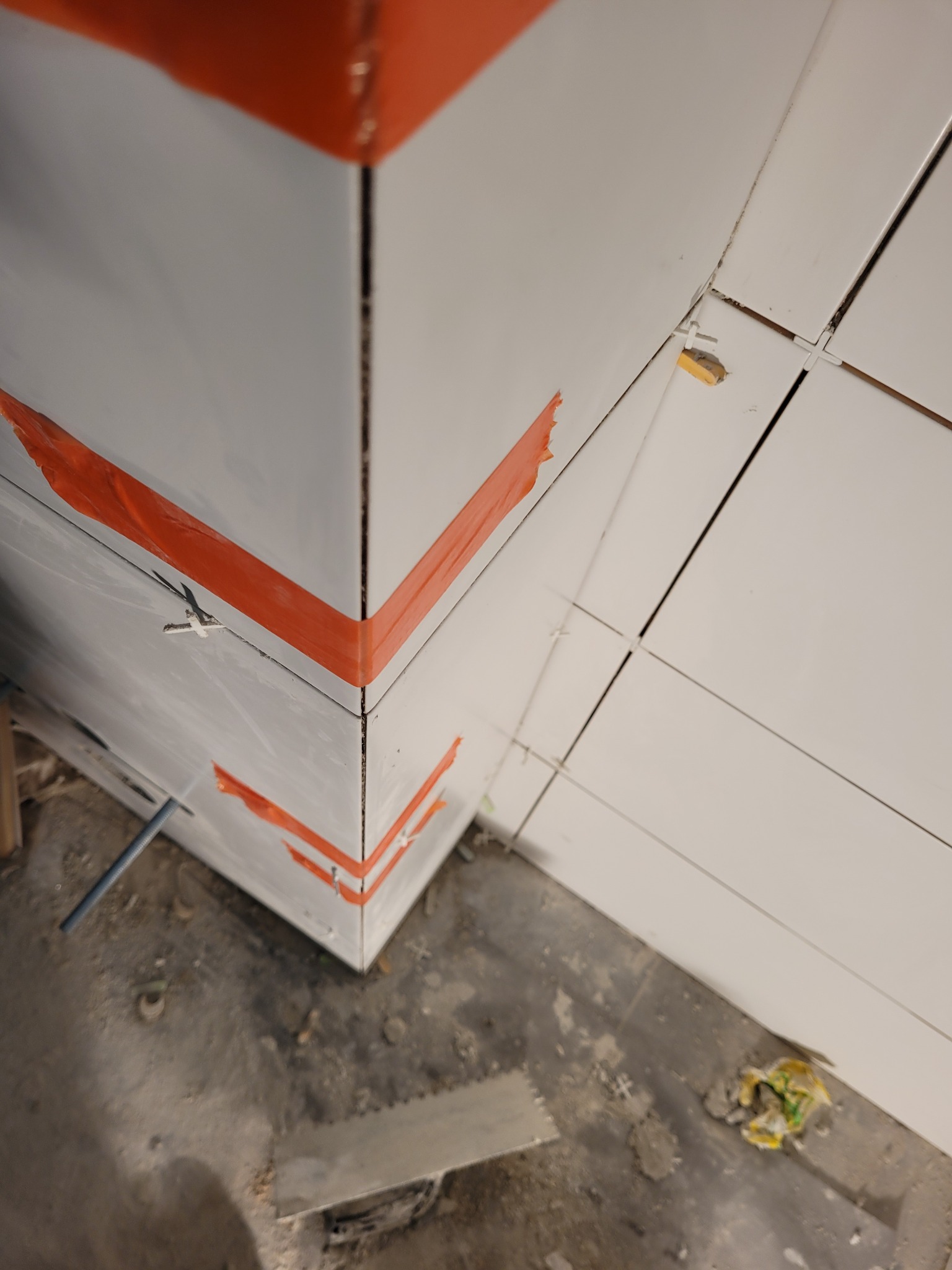 Narożnik ściany wyłożony białymi płytkami, zabezpieczony pomarańczową taśmą malarską, widoczne krzyżyki dystansowe i zabrudzony cementem szary podkład podłogowy.