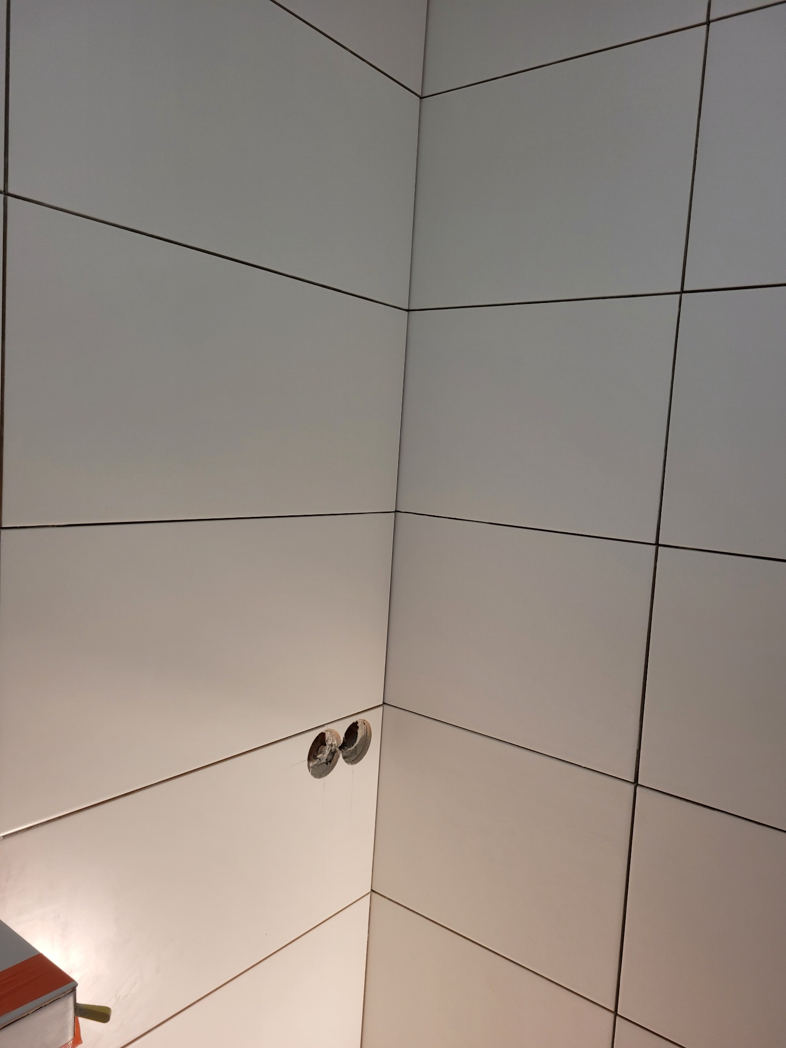 Ściana w rogu pomieszczenia wyłożona białymi, prostokątnymi płytkami z ciemnymi fugami. Widoczne dwa otwory pod instalację elektryczną, z których wycieka woda. W lewym dolnym rogu widoczny fragment...