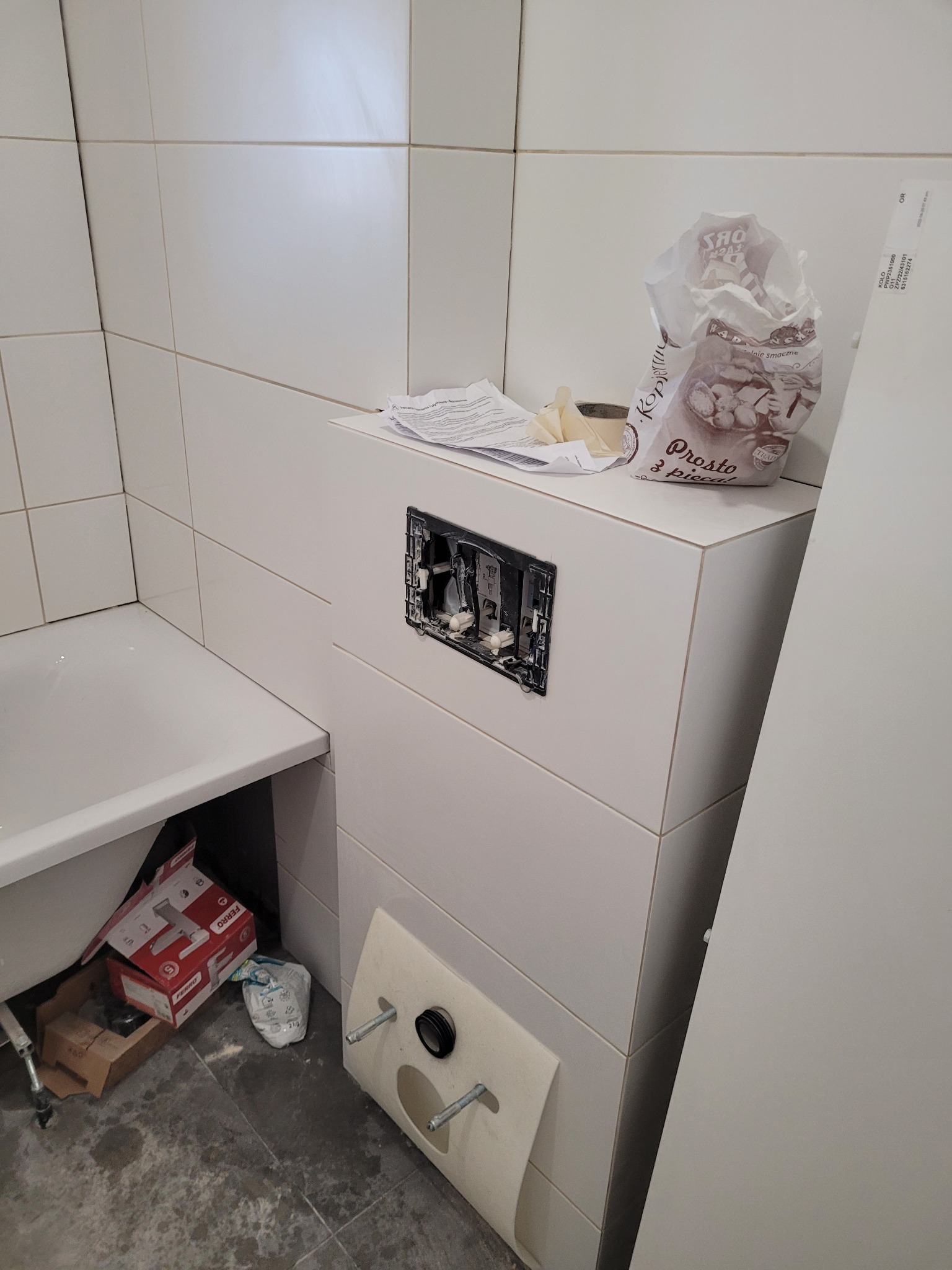 Biała łazienka w trakcie remontu, widoczna wanna, zabudowa stelaża podtynkowego z niedokończonym otworem na przycisk spłukujący, białe płytki na ścianach, na górze zabudowy leżą dokumenty...