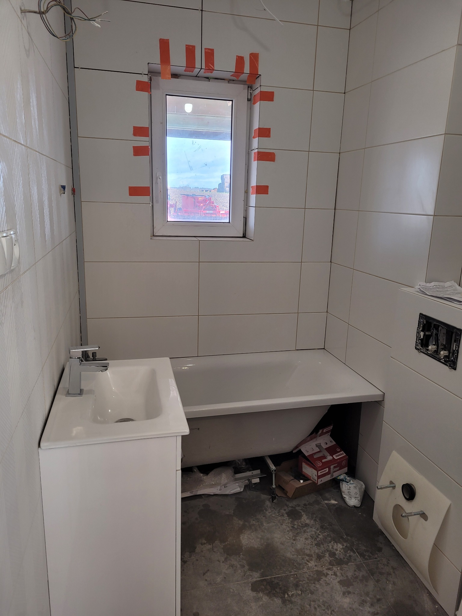 Wnętrze łazienki w trakcie wykończenia: białe płytki ścienne, okno zabezpieczone taśmą malarską, biała umywalka z szafką, wanna, widoczne elementy instalacji i materiały budowlane na podłodze.