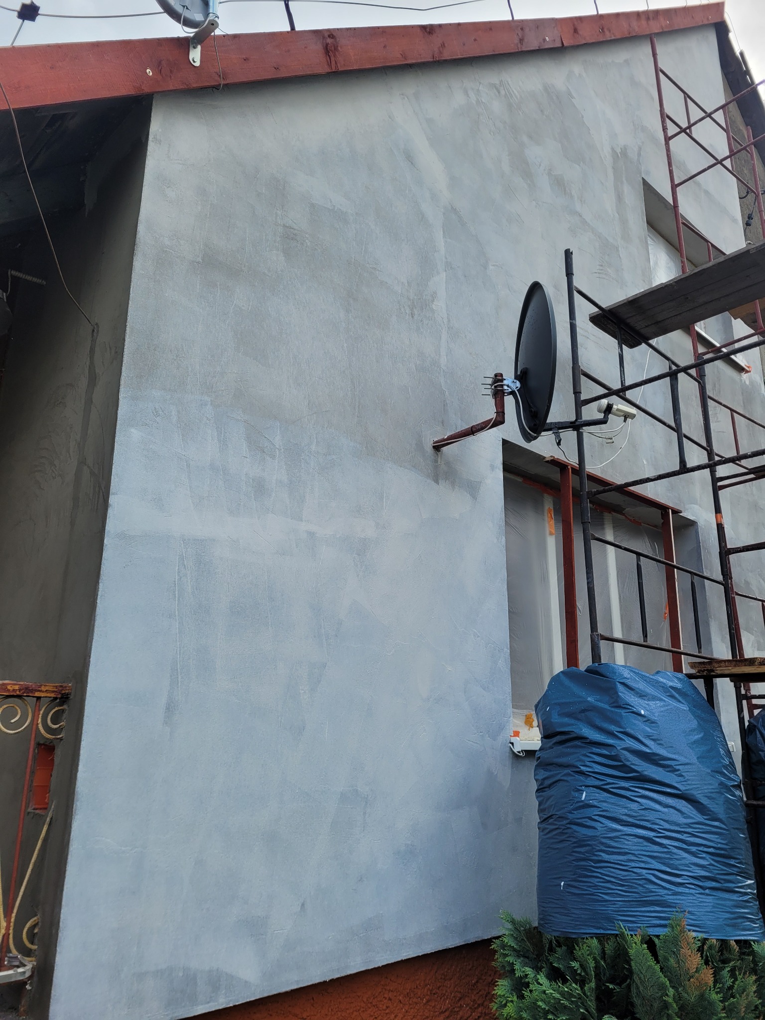 Ściana domu w trakcie wykonywania elewacji, widoczne rusztowanie, okno zabezpieczone folią, antena satelitarna i worki z materiałami budowlanymi.