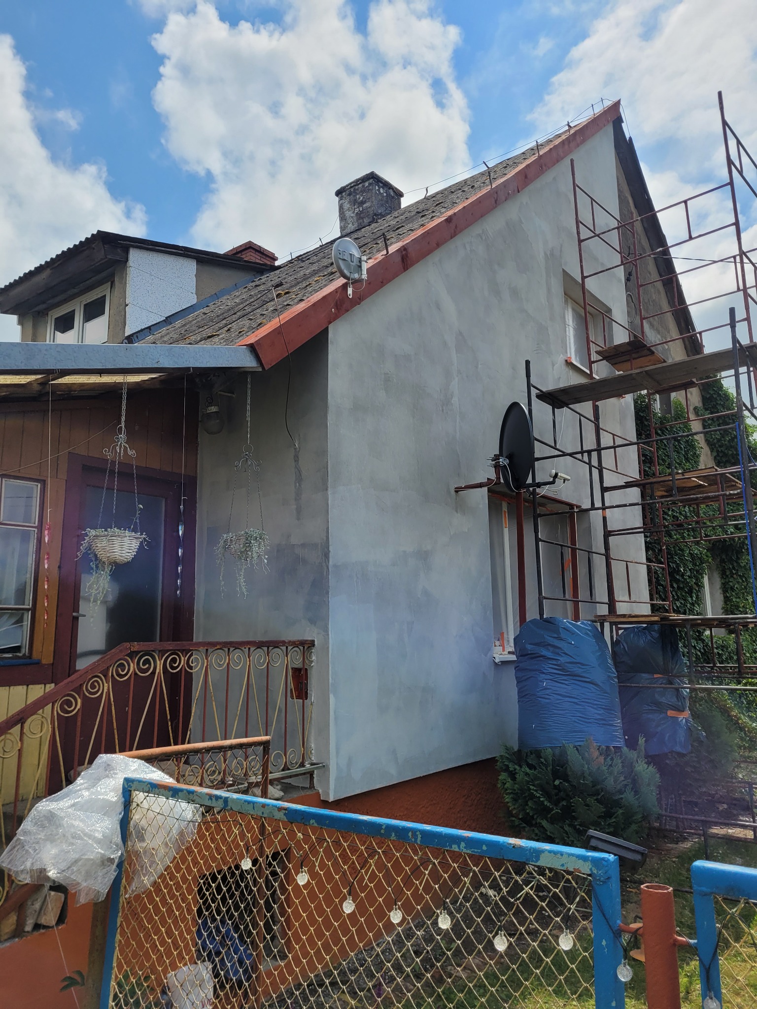 Dom w trakcie prac elewacyjnych, widoczne rusztowania, ściana po nałożeniu szarego tynku, antena satelitarna na ścianie, worki z odpadami budowlanymi u podnóża ściany.