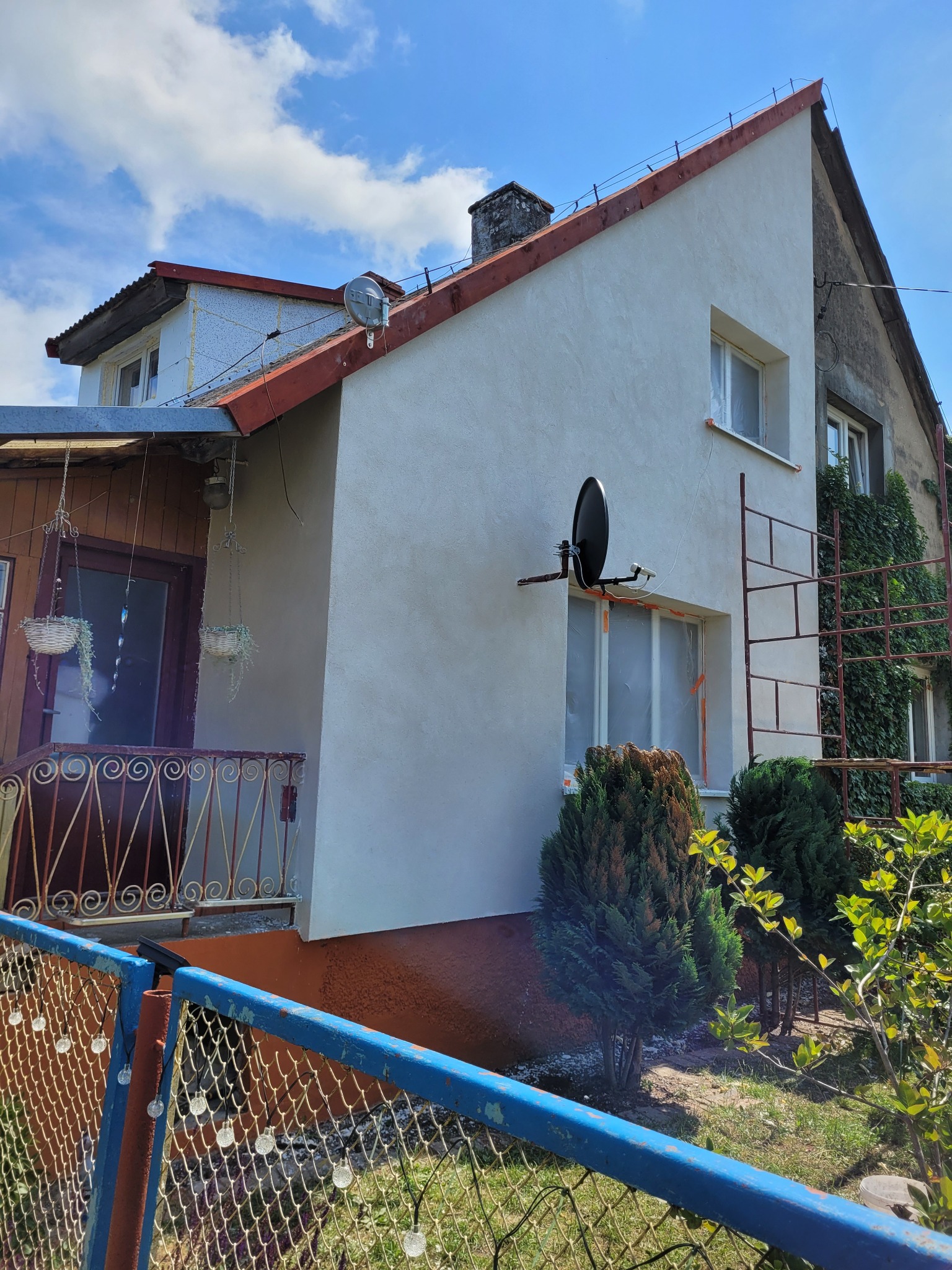 Odnowiona elewacja domu z zamontowaną anteną satelitarną, okna zabezpieczone folią malarską, drabina oparta o ścianę, widoczne pnącza na sąsiedniej części budynku.