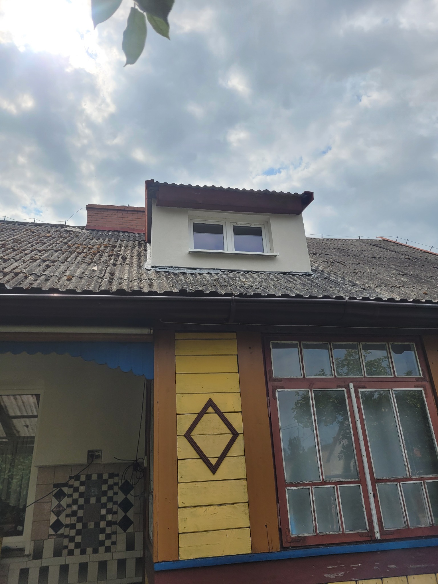 Fragment elewacji domu z oknem mansardowym, widoczne stare dachówki, drewniana weranda z wzorem rombu na żółtej desce.