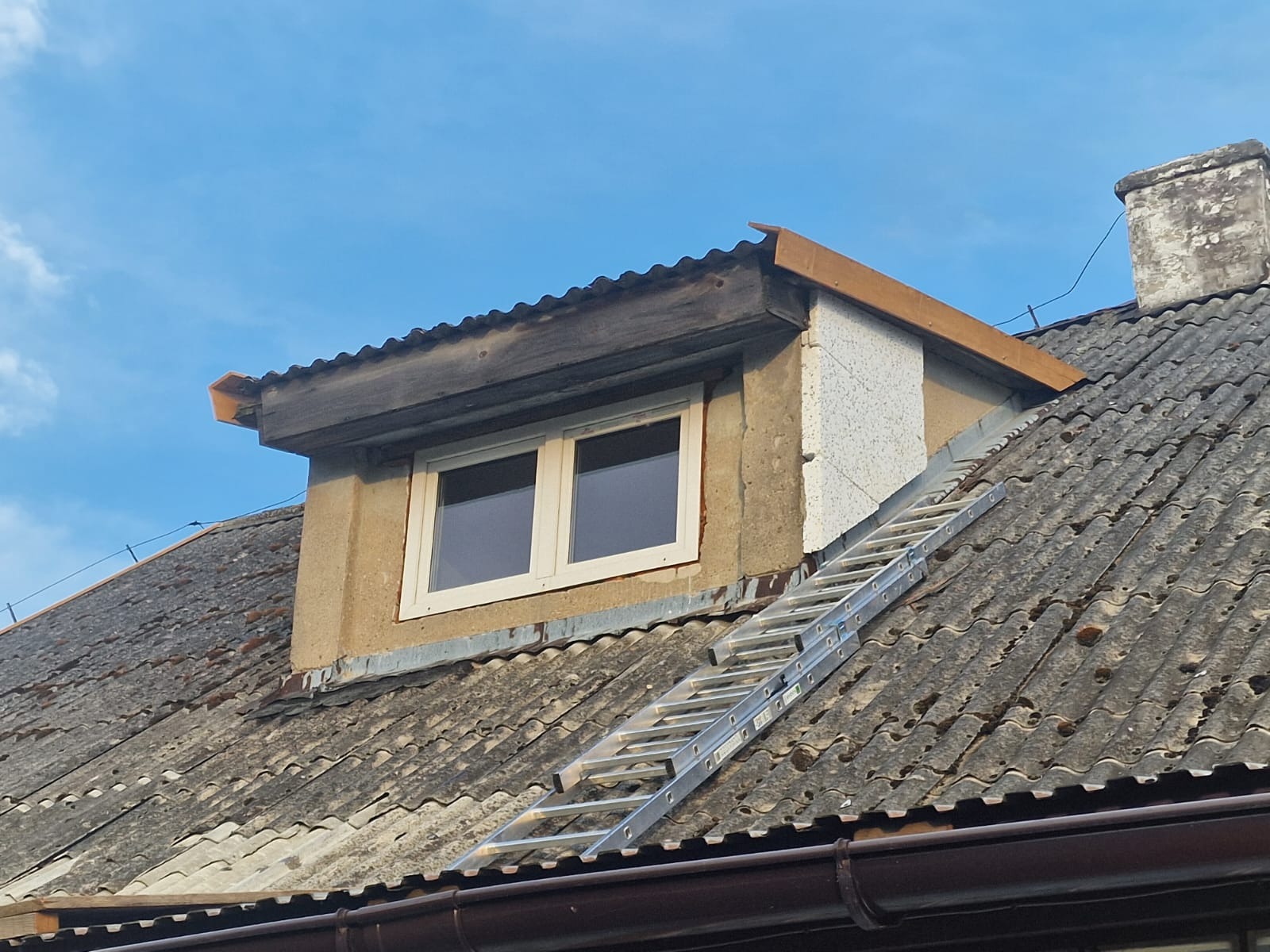 Stara dachówka z mchem, okno dachowe z drewnianą nadbudową, komin i drabina oparta o dach.