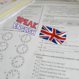 Otwarty podręcznik do nauki języka angielskiego z naklejką 'Speak English' i flagą Wielkiej Brytanii, uzupełniany przez ucznia, z widocznymi zadaniami gramatycznymi i zegarami do dopasowania.