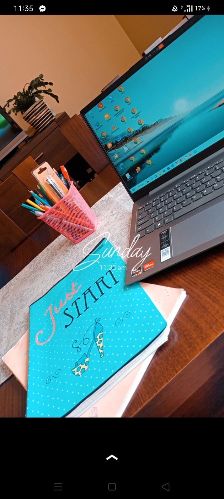 Laptop na biurku obok notesu z napisem 'Just START' i rysunkiem szpilki, w tle różowy pojemnik z długopisami i ołówkami, oraz roślina doniczkowa.