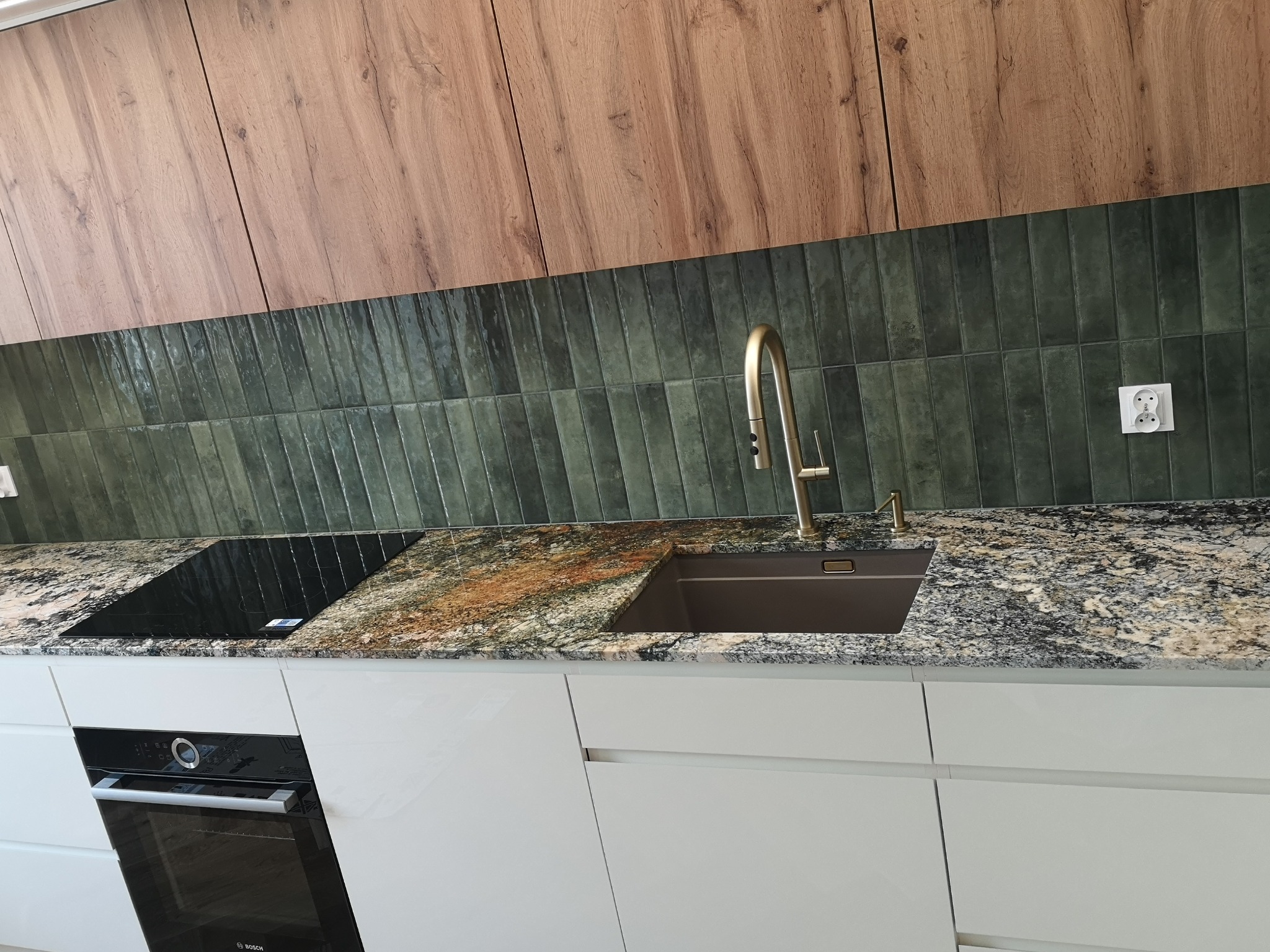 Kuchnia z blatem z kamienia o unikalnym wzorze, zielone płytki na ścianie i złota bateria. Nowoczesny design.